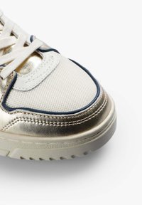 Primer plano de una zapatilla blanca con detalles metálicos dorados, tela de malla y ribete azul en la suela de goma texturizada.