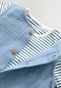Weicher blauer ärmelloser Baby-Body mit Holzknöpfen, der über einem weißen und navyblauen gestreiften Langarm-Baby-Body mit Druckknöpfen getragen wird.