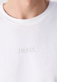 Gros plan d'une personne portant une chemise blanche avec le mot "BOSS" imprimé en lettres blanches sur la poitrine.