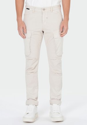 Beige, eng anliegende Cargohosen mit großen Seitentaschen, hochgekrempelt an den Knöcheln, kombiniert mit weißen Sneakers und einem weißen Hemd.