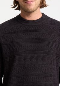 Sort tekstureret strikket sweater med ribbet rund hals, mønstrede sektioner og varierende overfladeteksturer over det hele.