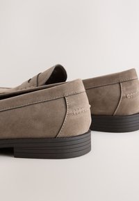 Paar taupefarbene Wildleder-Slipper mit schwarzen geriffelten Sohlen, aus einer niedrigen hinteren Perspektive vor einem einfachen hellen Hintergrund gezeigt.