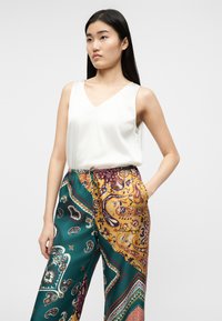 Femme aux longs cheveux noirs portant un haut blanc sans manches et un pantalon large multicolore à motifs cachemire et géométriques.