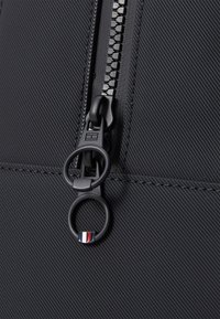 Tissu noir texturé avec une fermeture éclair proéminente dotée de deux tirettes circulaires. Une tirette présente un accent rayé en rouge, blanc et bleu.