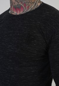 Fekete, texturált póló kerek nyakkivágással és rövid ujjakkal. Bal mellen egy kis fekete hímzett logó található.