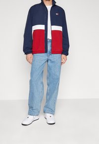 Granatowo-czerwona kurtka przeciwwiatrowa z białym poziomym paskiem, elastycznymi mankietami i ściągaczem u dołu. W połączeniu z jasnoniebieskimi dżinsami i białymi trampkami.