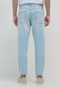 Uomo che indossa jeans in denim azzurro chiaro e scarpe casual beige, in piedi con la schiena rivolta verso la fotocamera su uno sfondo semplice.