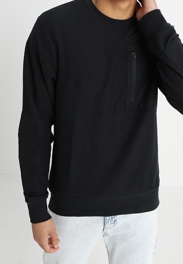 Sudadera negra de manga larga con tela texturizada, un bolsillo en el pecho con cremallera, y puños y dobladillo de canalé. Llevada con unos vaqueros de denim claro.