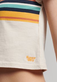 Beige T-shirt met korte mouwen, voorzien van veelkleurige horizontale strepen en een oranje "Super Dry" logo linksonder. Zachte katoenstructuur.