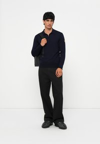 Polo de manga larga color navy con cuello de media cremallera, combinado con pantalones negros y zapatillas oscuras, llevando una mochila negra.