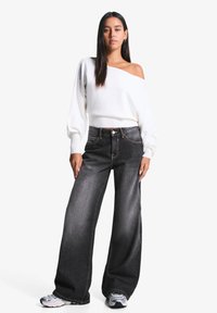 Bijela off-shoulder sweatshirt s balon rukavima, u kombinaciji s širokim tamnoplavim trapericama koje imaju kontrastne šavove i kopčanje na gumbi.