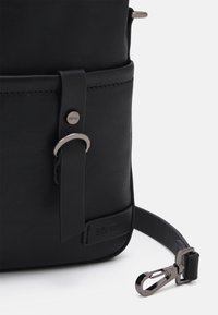 Roh černé kožené tašky s popruhem, na kterém je kovová spona a na spodní části vyražené logo Esprit, na bílém pozadí.