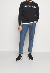 Man i svart Nike Air sweatshirt, blå uppvikta jeans, vita strumpor och svarta Nike sneakers som står mot en neutral bakgrund.