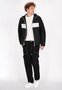 Svart jacka med vitt horisontellt accentpanel, ihopparad med svarta cargo-byxor med vit söm och fickor. Vita sneakers.