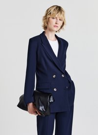 Blazer doppiopetto blu navy con bottoni dorati, ampie rever, e tasche, abbinato a una clutch in pelle nera e una camicia bianca.