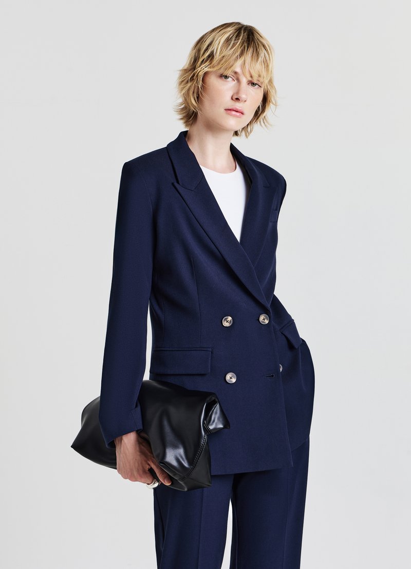 Blazer doppiopetto blu navy con bottoni dorati, ampie rever, e tasche, abbinato a una clutch in pelle nera e una camicia bianca.