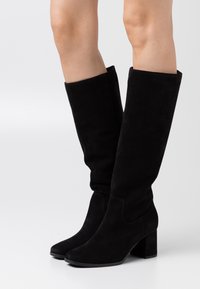 Botas altas de suede negro con un diseño ligeramente holgado, punta cuadrada y tacón grueso. Superficie lisa con detalles de costura.