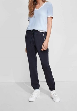 Vrouw draagt een lichtblauw V-hals shirt met korte mouwen, marineblauwe joggerbroek met ritszakken, witte sokken en witte sneakers, staand tegen een effen achtergrond.