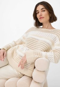 Maglione lavorato a maglia color crema con strisce orizzontali e dettagli in pizzo. Presenta polsini a coste e una vestibilità rilassata. Indossato sopra pantaloni beige.