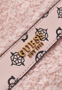 Tissu rose texturé avec une lanière en cuir rose ornée de motifs floraux noirs et de l'inscription dorée « GUESS EST 1981 ».