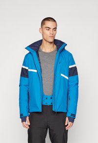 Blaue isolierte Jacke mit hohem Kragen, weißen und marineblauen Akzenten, mit Reißverschluss versehenen Taschen und leicht gekürztem Design. Getragen über einem grauen Oberteil.