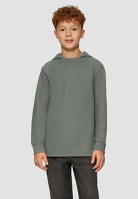 Ragazzo con capelli ricci che indossa una maglia a maniche lunghe con cappuccio di un verde spento e pantaloni grigio scuro, in piedi di fronte a uno sfondo grigio chiaro.