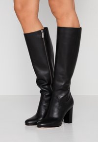 Nohy oblečené v černých kožených vysokých botách s blokovými podpatky a viditelnými bočními zipy, stojící na hladkém bílém povrchu.