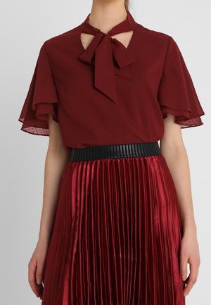 Vrouw draagt een bordeauxroze blouse met zwierige mouwen en een strik in de hals, gecombineerd met een glanzende geplooide bordeauxrode rok en een zwarte tailleband.