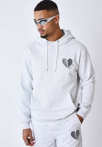 Sudadera con capucha gris, con un bolsillo central y un gráfico de un corazón roto. Tejido texturizado, con prendas inferiores grises a juego.