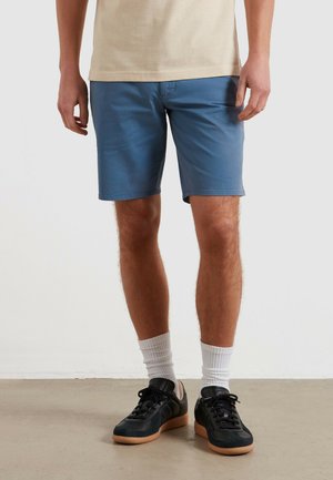 Uomo che indossa pantaloncini blu fino al ginocchio, calzini bianchi alti, sneakers nere e una camicia beige, in piedi su un pavimento semplice su sfondo bianco.