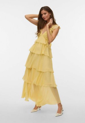 Žena pózujúca v vrstvených, žltých bezrukávových maxi šatách s volánikmi a bielych topánkach na špic na jednofarebnom pozadí.