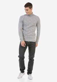 Grijze gebreide coltrui met een gestructureerd blokpatroon, gecombineerd met zwarte jeans en witte sneakers. Model staat casual.