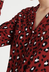 Blouse à motifs léopard rouge avec des taches noires et blanches, dotée d'un col et de manches longues, fabriquée dans un tissu lisse.