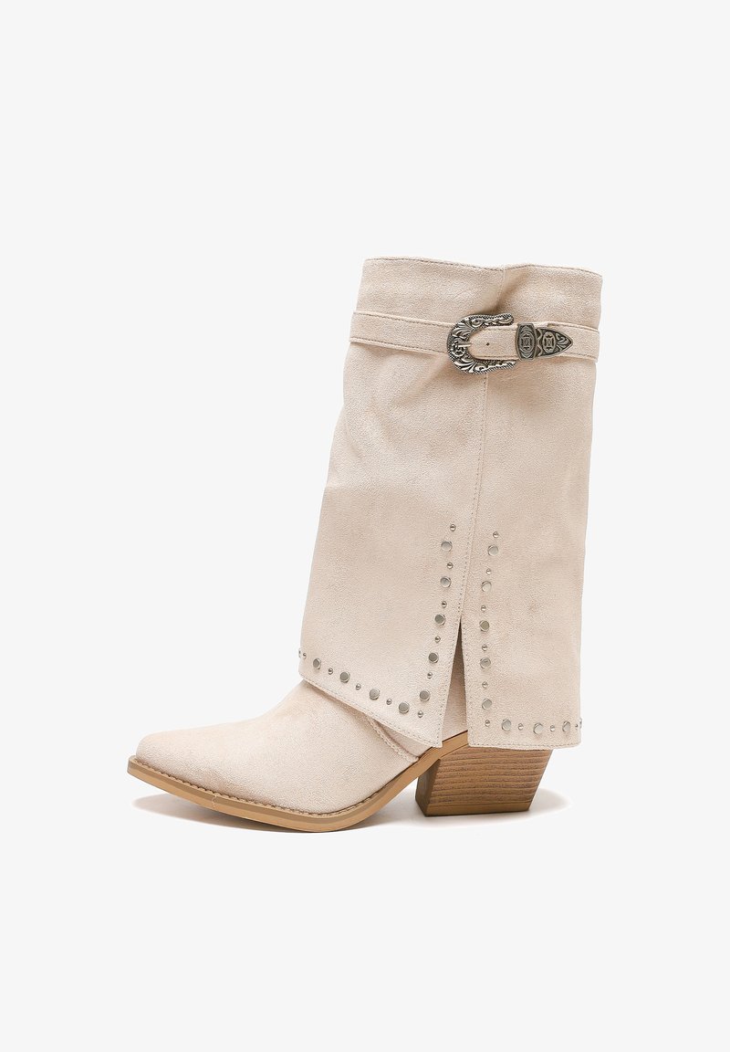 Botín de gamuza beige con punta afilada, acentos con tachuelas plateadas, una hebilla decorativa y un tacón de madera apilada.