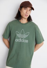 Zielona bawełniana koszulka z białym logo Adidas w kształcie trifolium. Okrągły dekolt, krótkie rękawy, miękka faktura, luźny krój.