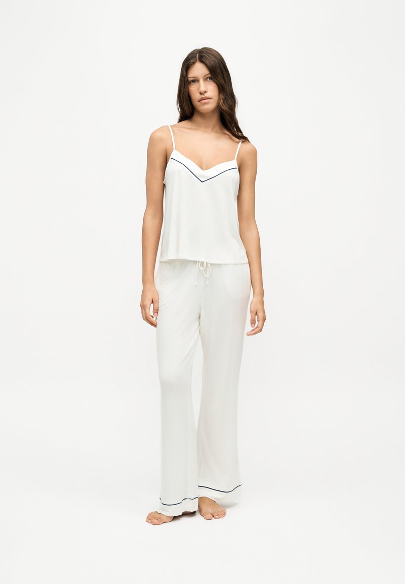 Femme debout pieds nus, portant un haut blanc sans manches et un pantalon blanc ample avec un liseré foncé au niveau de l'encolure et des revers de pantalon.