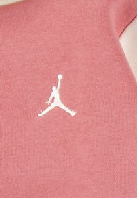 Roze tkanina s mekim teksturom i bijelim izvezenim Jumpman logom. Dizajn je jednostavan bez dodatnih uzoraka ili akcenata.