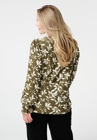 Blouse vert olive avec un motif floral blanc, manches bouffantes et détails froncés aux poignets. Tissu texturé et ourlet arrondi.