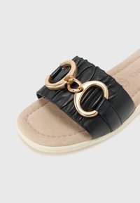 Black slide sandal made of textured synthetic material, featuring two interlocking gold rings and a beige cushioned footbed.
