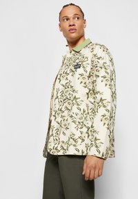Floral patterned jacket in cream, featuring green collar and embroidered logo. Lightweight fabric with a relaxed fit and side pockets.