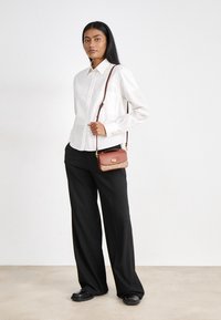 Barna bőr crossbody táska mintás aljjal, bronz színű fémalkatrészekkel és állítható pánttal. Fehér csíkos inggel és fekete nadrággal párosítva.