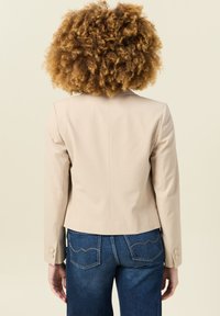 Blazer beige su misura con un design aderente, dotato di una cucitura sul retro e dettagli con bottoni sulle maniche, abbinato a jeans blu scuro.