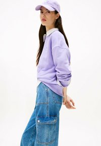 Sudadera lila con puños acanalados, superpuesta a una camisa blanca con cuello. Combinada con pantalones cargo de denim azul holgados que cuentan con bolsillos laterales.