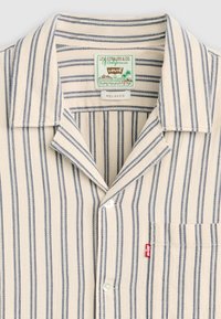 Beige kortermet skjorte med blå vertikale striper, krage og brystlomme. Har en Levi's-merkelapp og knappelukking.