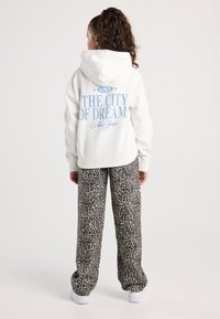 Giovane con capelli ricci indossa un hoodie bianco con la scritta "USA La Città dei Sogni New York", pantaloni a stampa leopardata e sneaker bianche, in piedi di spalle.