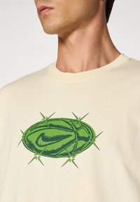 Person iført en cremefarvet skjorte med et grønt grafik af en kaktuslignende oval med en stiliseret Nike swoosh på brystet.