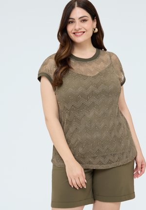 Femme aux longs cheveux bruns portant des boucles d'oreilles créoles dorées, un haut en dentelle à motif zigzag vert olive et un short assorti longueur genou, souriante avec une posture détendue.