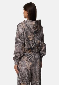 Camo hoodie met een mix van bruin, grijs en zwart, voorzien van een trekkoordkap, elastische manchetten en een relaxte pasvorm. De stof toont bladeren en schorspatronen.