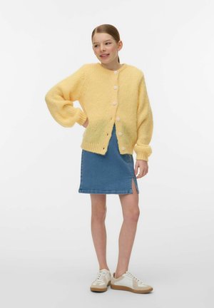 Junges Mädchen posiert in einem gelben, geknöpften Cardigan, blauem Jeansrock mit Schlitz und weißen Turnschuhen vor schlichtem Hintergrund.