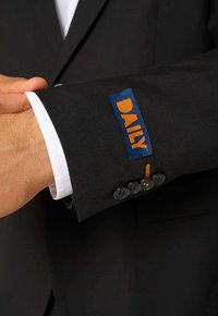 Rukáv čierneho saku s modrou nášivkou s nápisom "DAILY" v oranžovej farbe, s gombíkovými akcentmi, bielym manžetovým detailom a oranžovým prešívaním.